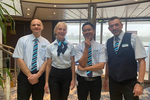 Quatre personnes souriantes en uniforme avec des cravates rayées bleues et noires se tiennent ensemble dans une pièce bien éclairée avec de grandes fenêtres. La pièce a un design moderne avec des panneaux en bois et une plante visible sur le côté gauche.